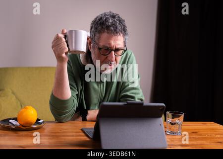 Senior man with glasses using a tablet while drinking coffee at home in a calm morning routine. Stock Photo