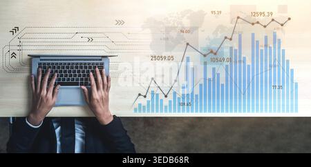 A businessman works on a laptop surrounded by financial data visualizations and charts, depicting market trends and analytics on a wooden desk surface Stock Photo
