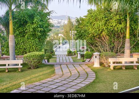 A well-manicured tropical park featuring a curved stone pathway with grass growing between rectangular pavers, leading downhill through lush Stock Photo