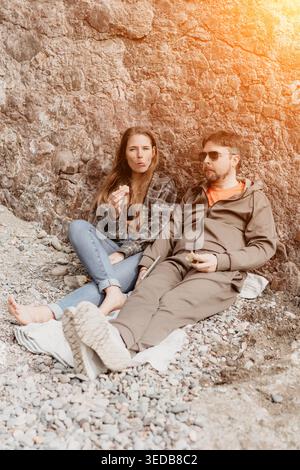 Couple beach picnic enjoying snacks while sitting on rocky coastline near a rock wall under sunlight Stock Photo