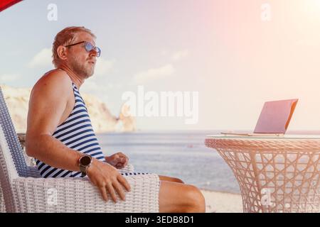 Man laptop beach sitting at a seaside terrace working remotely during a summer vacation while enjoying the ocean view Stock Photo