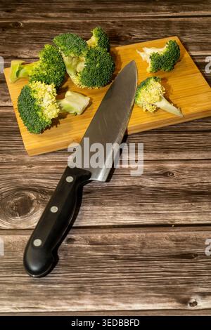 Broccoli florets are arranged on a wooden cutting board alongside a chef's knife, set against a rustic wooden backdrop. Stock Photo
