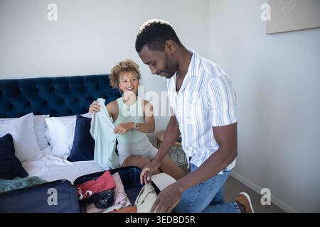 Diverse couple sitting on bed edge holding striped shirt and packing suitcase in bedroom Stock Photo