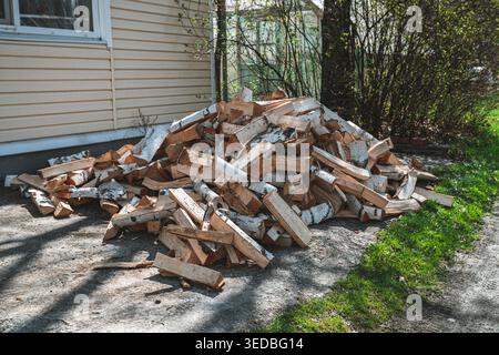 Large pile of chopped birch firewood logs, with white bark visible, lying on the ground next to a building exterior wall and green grass, representing Stock Photo
