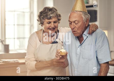 Happy, old couple and blow candle on birthday for celebration, milestone event and cupcake. Hug, senior man and party hat with partner in home for Stock Photo