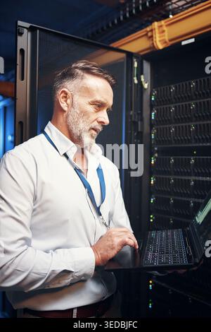 Technician, man and typing on laptop in server room for maintenance, diagnostics and IT solution. Engineer, mature person and tech in data center for Stock Photo
