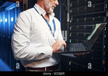 Hands, man and typing on laptop in data center for maintenance, diagnostics and IT support. Technician, mature person and tech in server room for Stock Photo