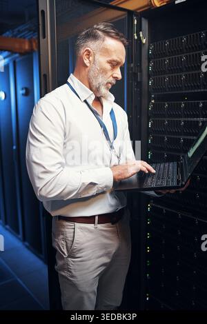 Engineer, man and typing on laptop in server room for maintenance, diagnostics and IT support. Technician, mature person and tech in data center for Stock Photo