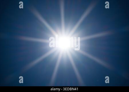 A symmetrical sunstar glows brightly in the center of a dark blue sky. The clean composition highlights natural light rays and sunbeams Stock Photo