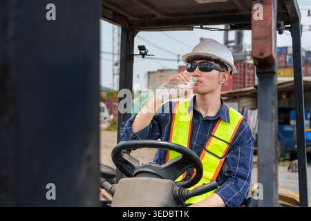 Young Asian male worker hydrates in a forklift at a shipping yard for safety and wellness in the logistics and transport industry in Asia. Stock Photo
