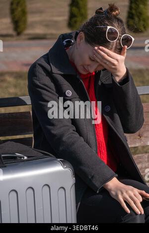 Woman sitting on an outdoor bench with a suitcase, covering her face with her hand due to a headache and feeling overwhelmed by travel stress and exha Stock Photo