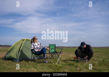Young man and woman spending leisure time together camping, preparing for an outdoor meal with a portable barbecue next to a green tent on a grassy fi Stock Photo