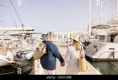 Senior couple enjoying summer vacation stroll, holding hands at a sunny pier with boats Stock Photo