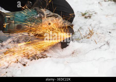 Worker uses an angle grinder to cut metal outdoors, producing bright orange sparks that scatter across the snowy ground. Cold-weather construction wor Stock Photo