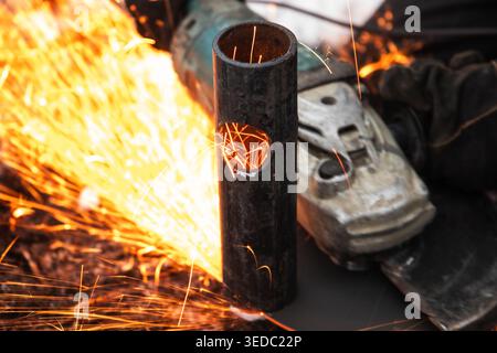 Worker using an angle grinder to cut a steel pipe, sending bright orange sparks across an industrial workshop. Image conveys heat, power, precision, m Stock Photo