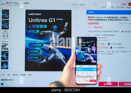 ZHONGSHAN China-May 4 2026: hand holding a smartphone displaying the Unitree G1 humanoid robot product page while a monitor shows the same product lis Stock Photo