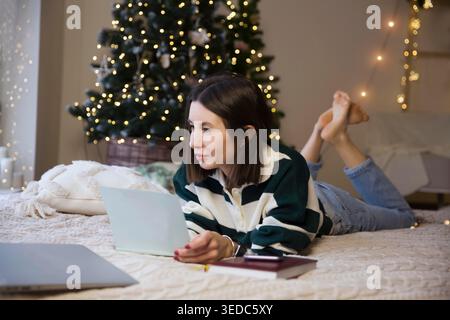 Woman writes checklist of plans and dreams for next year lying on the bed of a house in the Christmas interior. Wish list for Christmas. Gift expense planning. Goals Stock Photo