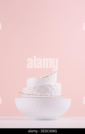White elegant plates in stack with thin golden bezel, bizarre texture on white wooden table, pastel pink wall, vertical. Modern home decor with elegant crockery. Stock Photo