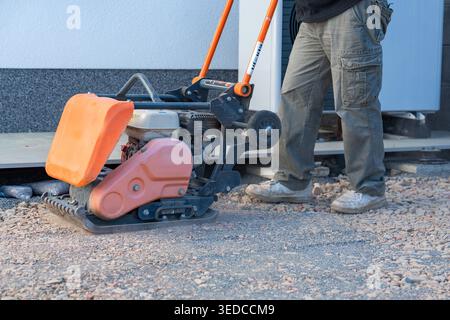 Worker uses a vibratory plate compactor for preparing a base layer. Essential step before paving, decking, or foundation installation. Stock Photo