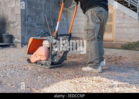Worker operates a vibratory plate compactor on a gravel surface at a construction site. The machine compacts the ground to create a stable base for Stock Photo