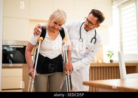 A healthcare professional assists a senior patient with mobility, guiding her carefully while she wears a back support brace and using crutches. Stock Photo