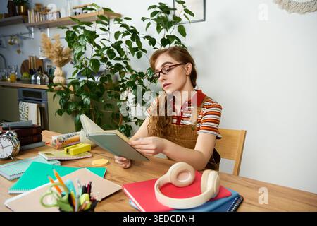 A young woman focuses on her studies while sitting at a wooden table filled with colorful notebooks. Stock Photo
