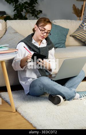Young student deeply engaged in her studies while seated comfortably at home surrounded by books. Stock Photo