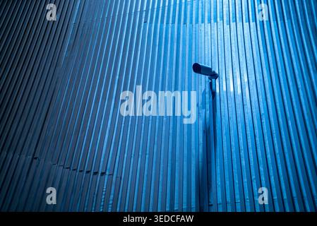 A surveillance camera mounted on a textured metal wall, illuminated by blue light, creates an intriguing visual of security and urban design elements. Stock Photo