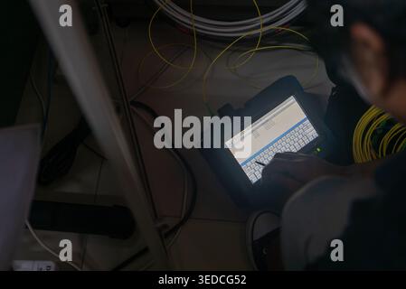 A network technician focused on a tablet device, handling cables in a complex workspace. This setup highlights technical skills and equipment in a mod Stock Photo