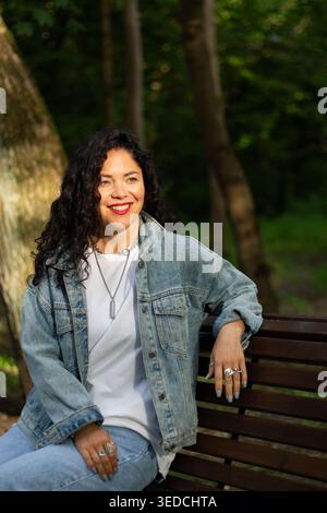 Joyful dark haired curly woman laughing in sunlit park. Young woman with beautiful dark curls smiles brightly being in park. Natural sunlight. Happy e Stock Photo