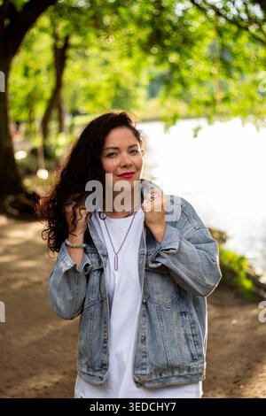 Woman relaxing in green park, smiling at camera. Casual and authentic moment of curly haired woman creating friendly and approachable vibe. Great for Stock Photo