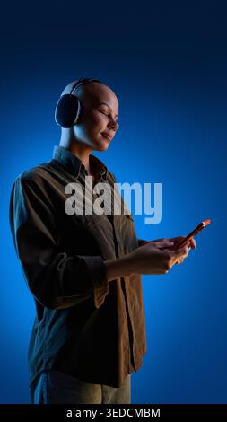Bald woman wearing headphones holding smartphone on blue gradient background. Stock Photo