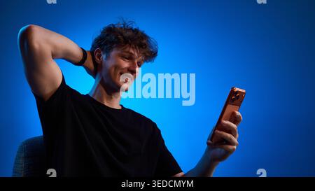 Smiling young man reading smartphone message in blue studio light Stock Photo