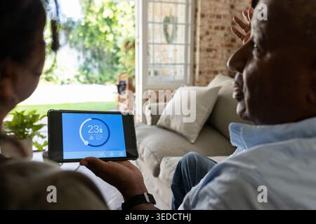 African senior couple viewing tablet thermostat 23Â°C while sitting on light sofa with garden view Stock Photo