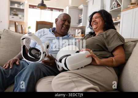 African American couple laughing, sitting on light sofa in open kitchen, holding white VR headsets Stock Photo