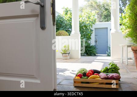 Wooden crate of produce is sitting on tiled porch near open white door, sunlight casting shadows Stock Photo