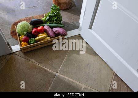Wooden crate is sitting on stone tiles near open white door, holding cabbage, eggplant, cucumbers Stock Photo