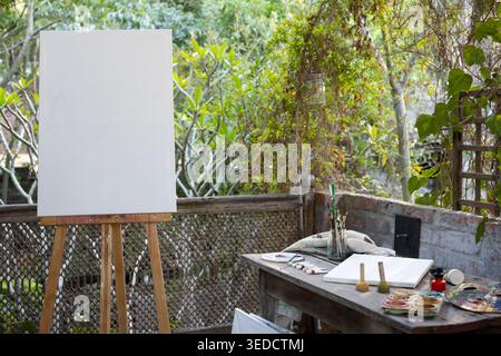 Primed canvas standing on easel on garden patio beside work table with brushes paints palette vines Stock Photo