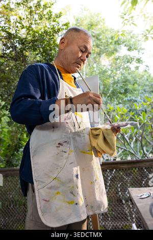 Senior Asian man standing on deck in paint apron, holding palette and brush, mixing paint Stock Photo