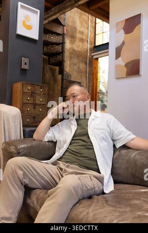 Senior Asian man sitting on brown leather sofa by metal spiral staircase, wearing olive green shirt Stock Photo