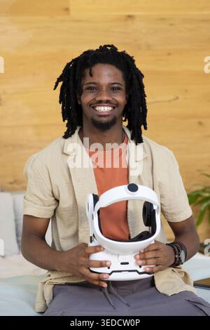 African American man smiling and sitting at home against wooden panel wall holding white VR headset Stock Photo