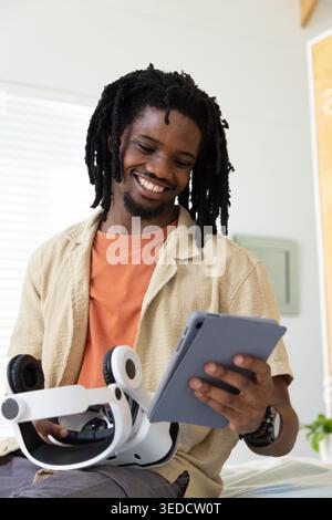 African man wearing beige overshirt sitting at home holding gray tablet and white VR headset Stock Photo