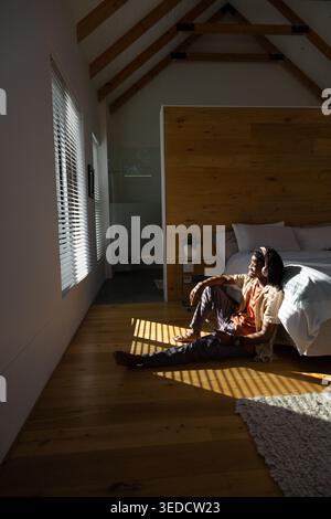 Adult African man sitting on wood floor next to bed holding smartphone, headphones on, copy space Stock Photo