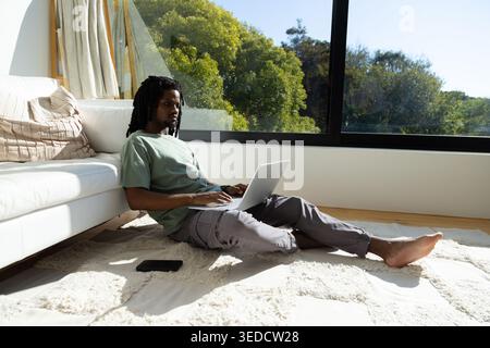 African American man sitting in living room on rug using silver laptop with black smartphone nearby Stock Photo
