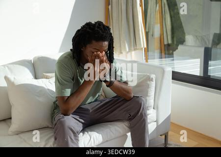 Adult African man sitting on white sofa covering face with hands by glass window, wearing bracelets Stock Photo