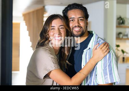 Diverse couple smiling and embracing while standing in modern kitchen near wooden slat divider Stock Photo