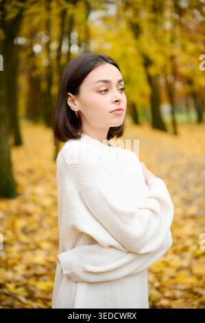 Young woman wrapped in white knitted sweater standing in fall park, glancing aside against golden foliage. Perfect for fashion, beauty, cozy lifestyle, seasonal branding, and advertising Stock Photo