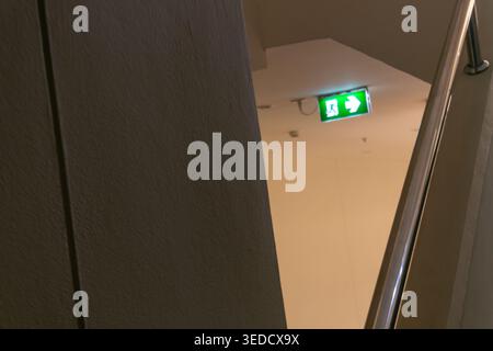 A close-up view of a glowing emergency exit sign illuminated on a wall near a staircase, showcasing safety features in a contemporary building environ Stock Photo