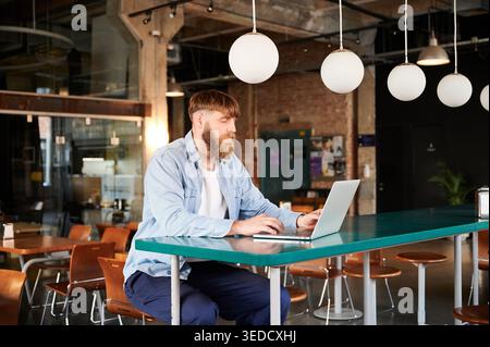 Freelancer with beard using notebook computer at high table in loft style workspace, suited for technology, entrepreneurship, online business, and work anywhere themes Stock Photo