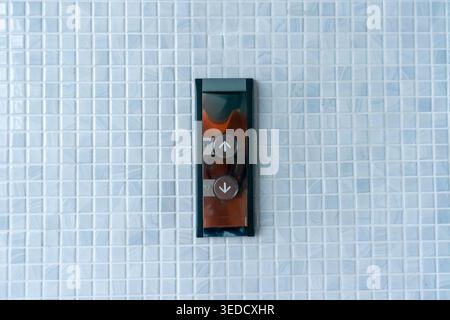 Close-up of an elevator control panel featuring up and down buttons on a textured blue mosaic wall. Ideal for themes of modern architecture and techno Stock Photo